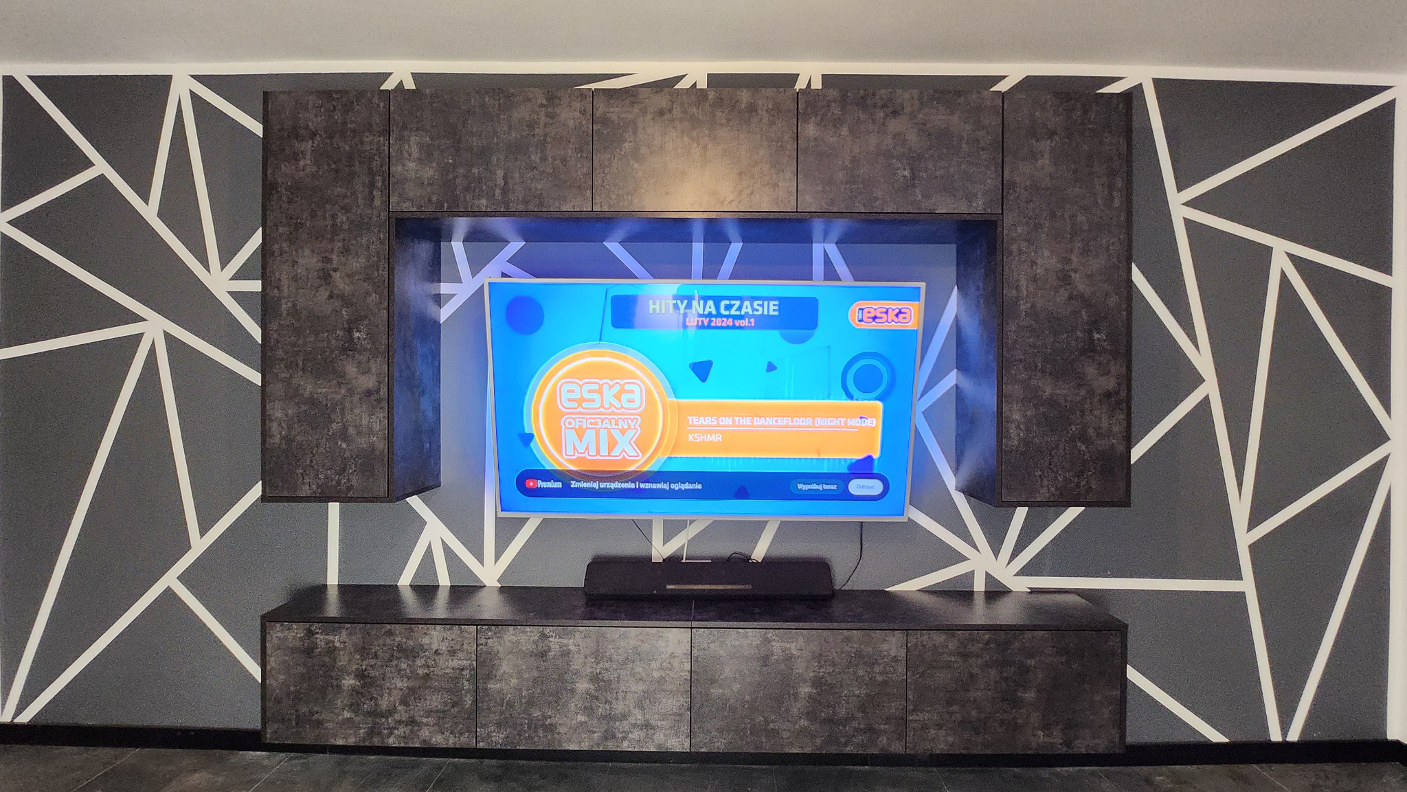 Ciemnoszara zabudowa meblowa z szafkami wiszącymi i dolną szafką RTV, na ścianie wzór geometryczny w odcieniach szarości i bieli, wnęka z telewizorem.