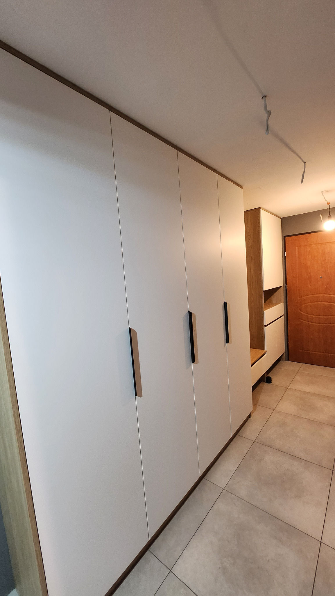 Biała szafa w zabudowie z ciemnymi, pionowymi uchwytami, oświetlenie sufitowe, płytki podłogowe w odcieniach szarości, widok na przedpokój z drewnianymi drzwiami wejściowymi.