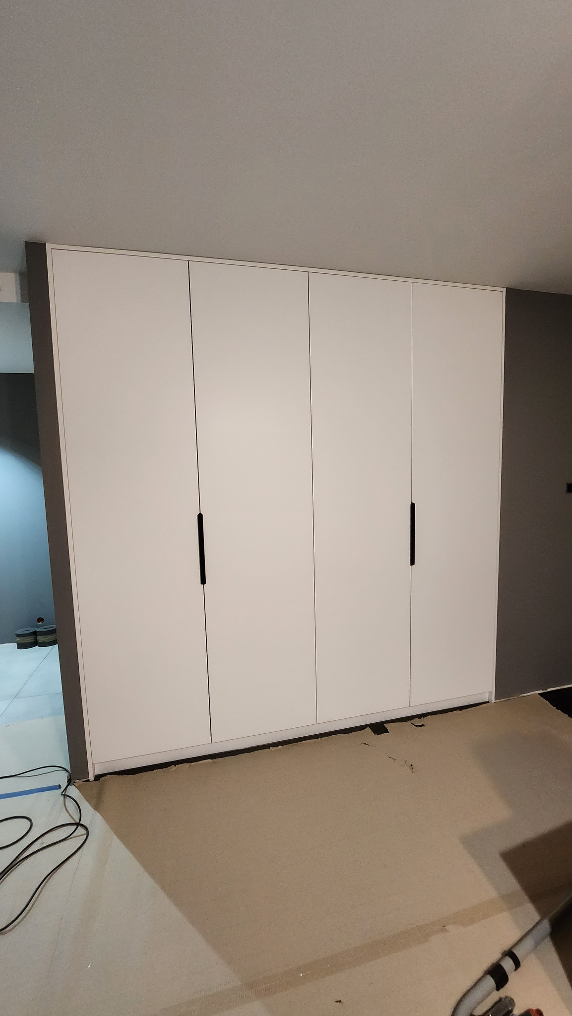 Biała, czterodrzwiowa szafa wnękowa z minimalistycznymi, czarnymi uchwytami, widoczna podczas montażu; podłoga zabezpieczona kartonem, widoczne kable i rura od odkurzacza.