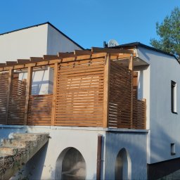 Arkadiusz Brożek - Drewniany taras z ażurową zabudową na piętrze domu z białymi ścianami i widocznymi schodami wejściowymi.