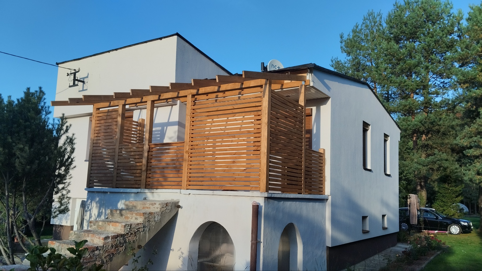 Drewniany taras z ażurową zabudową na piętrze domu z białymi ścianami i widocznymi schodami wejściowymi.