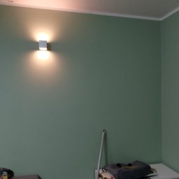 Krzysztof Marcińczyk - Ściana pomalowana na zielono z zamontowanym kinkietem, widoczny fragment mebla i akcesoria.