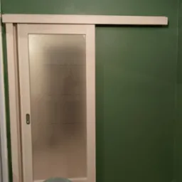 Zamontowane przesuwne drzwi z matowego szkła z poziomymi paskami na tle zielonej ściany, widoczny stołek monterski.