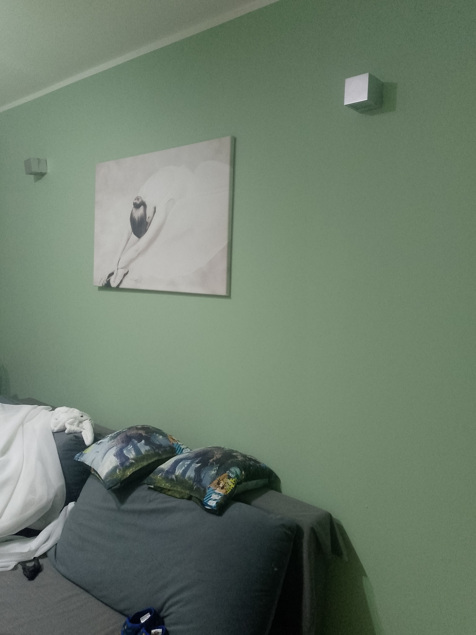 Ściana pomalowana na pistacjowy kolor z kwadratowym kinkietem i obrazem baletnicy nad szarą sofą z poduszkami.