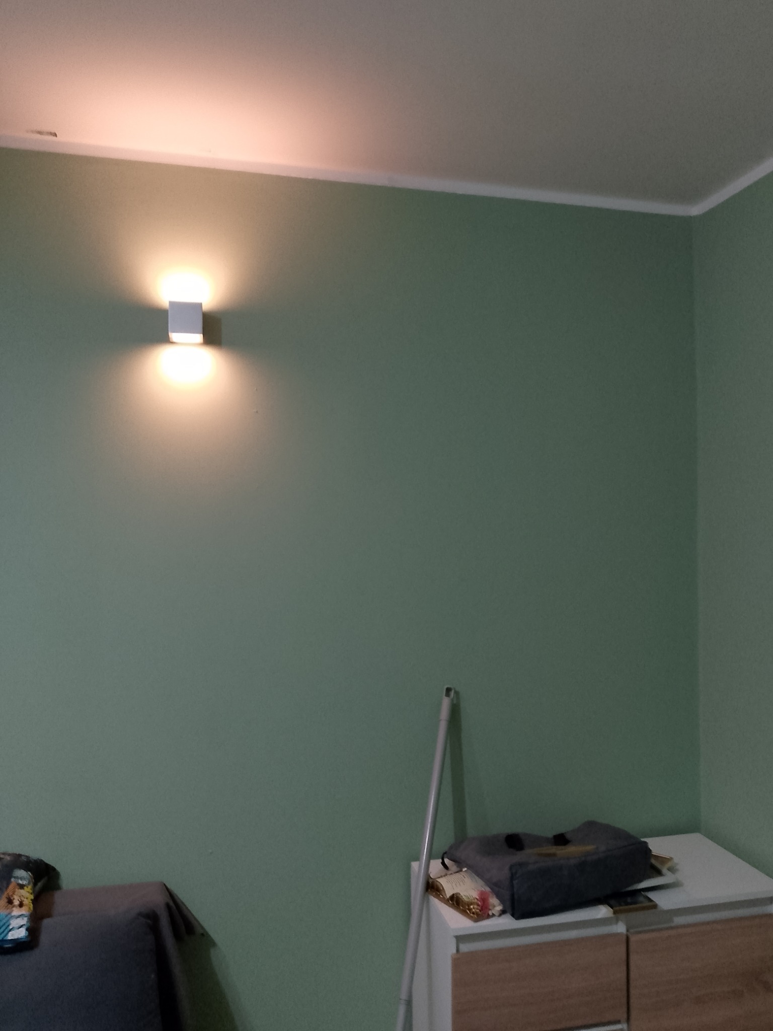 Ściana pomalowana na zielono z zamontowanym kinkietem, widoczny fragment mebla i akcesoria.