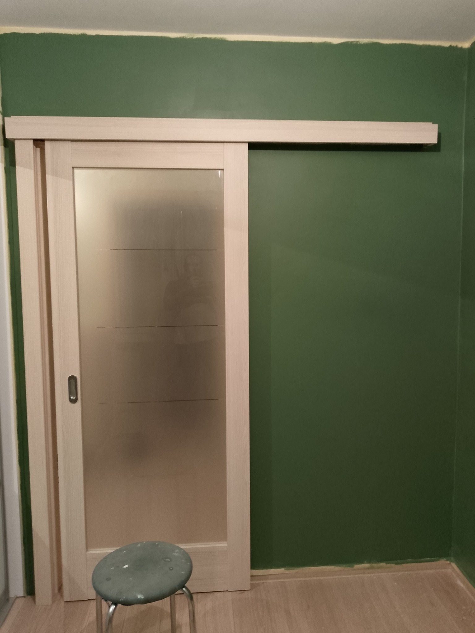 Zamontowane przesuwne drzwi z matowego szkła z poziomymi paskami na tle zielonej ściany, widoczny stołek monterski.