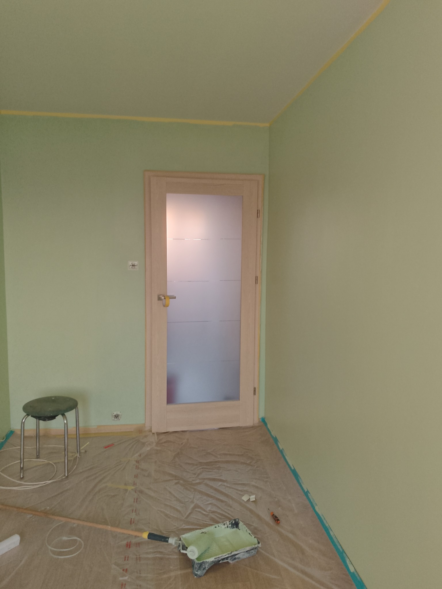 Pokój w trakcie malowania na pistacjowy kolor, zabezpieczona folią podłoga, widoczny wałek i kuweta malarska, taśma malarska przy suficie i podłodze, drzwi z matową szybą.