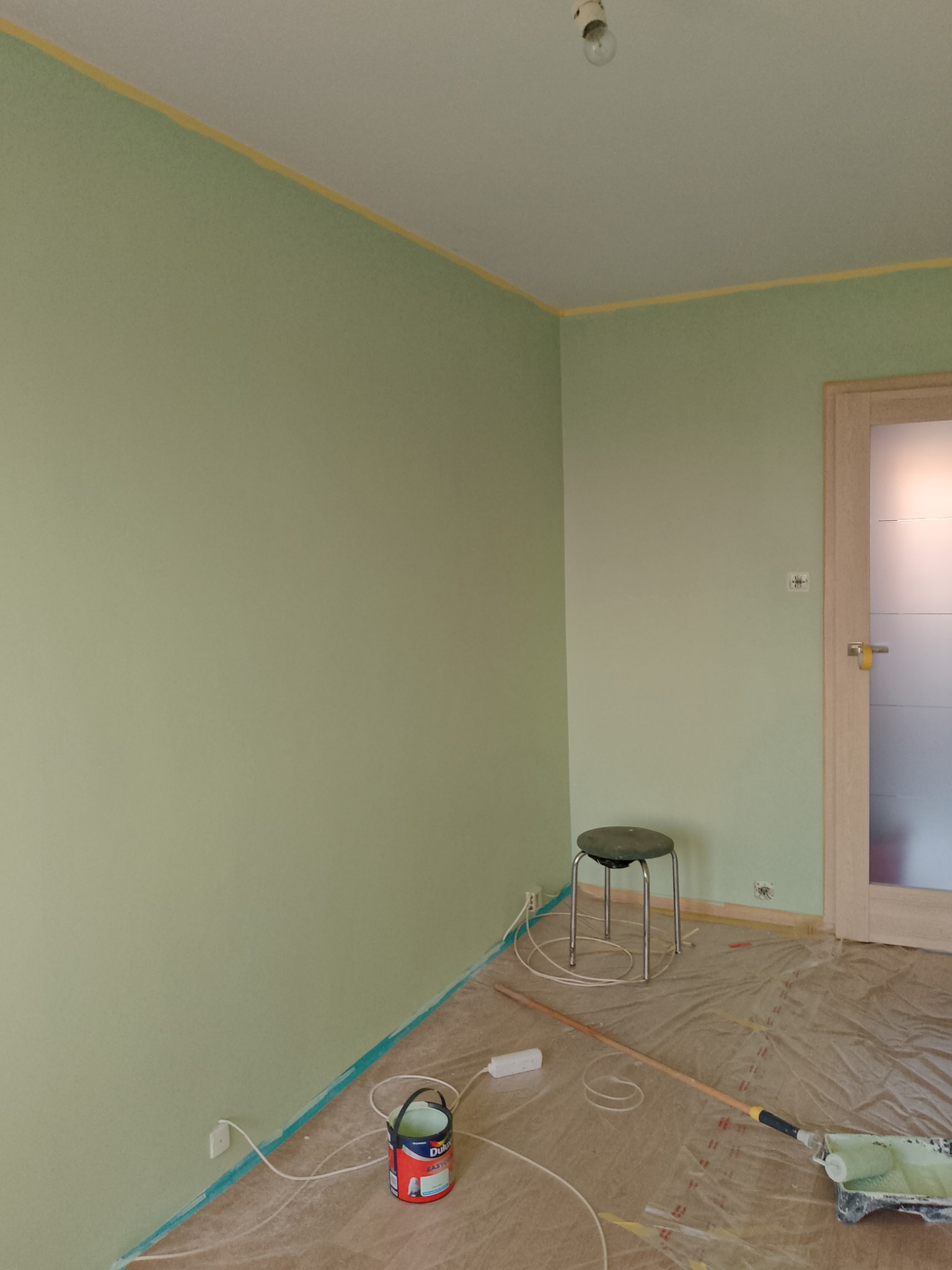 Pokój w trakcie malowania na jasnozielony kolor, zabezpieczony folią malarską, widoczna puszka z farbą Dulux, wałek i taboret.