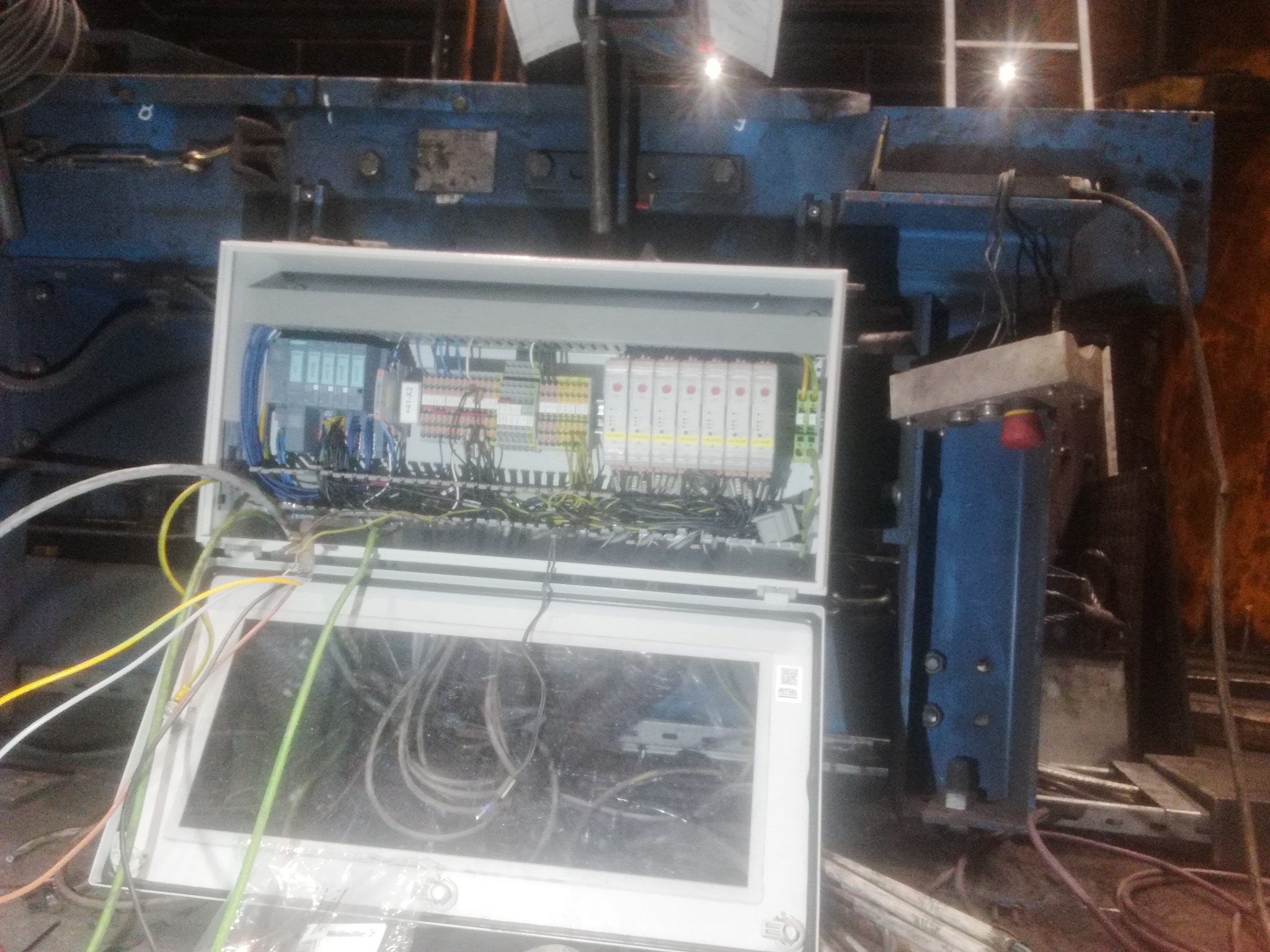 Otwarta skrzynka sterownicza z widocznymi elementami automatyki, przewodami i panelem operatorskim, umieszczona w przemysłowym otoczeniu z elementami maszyny w tle.