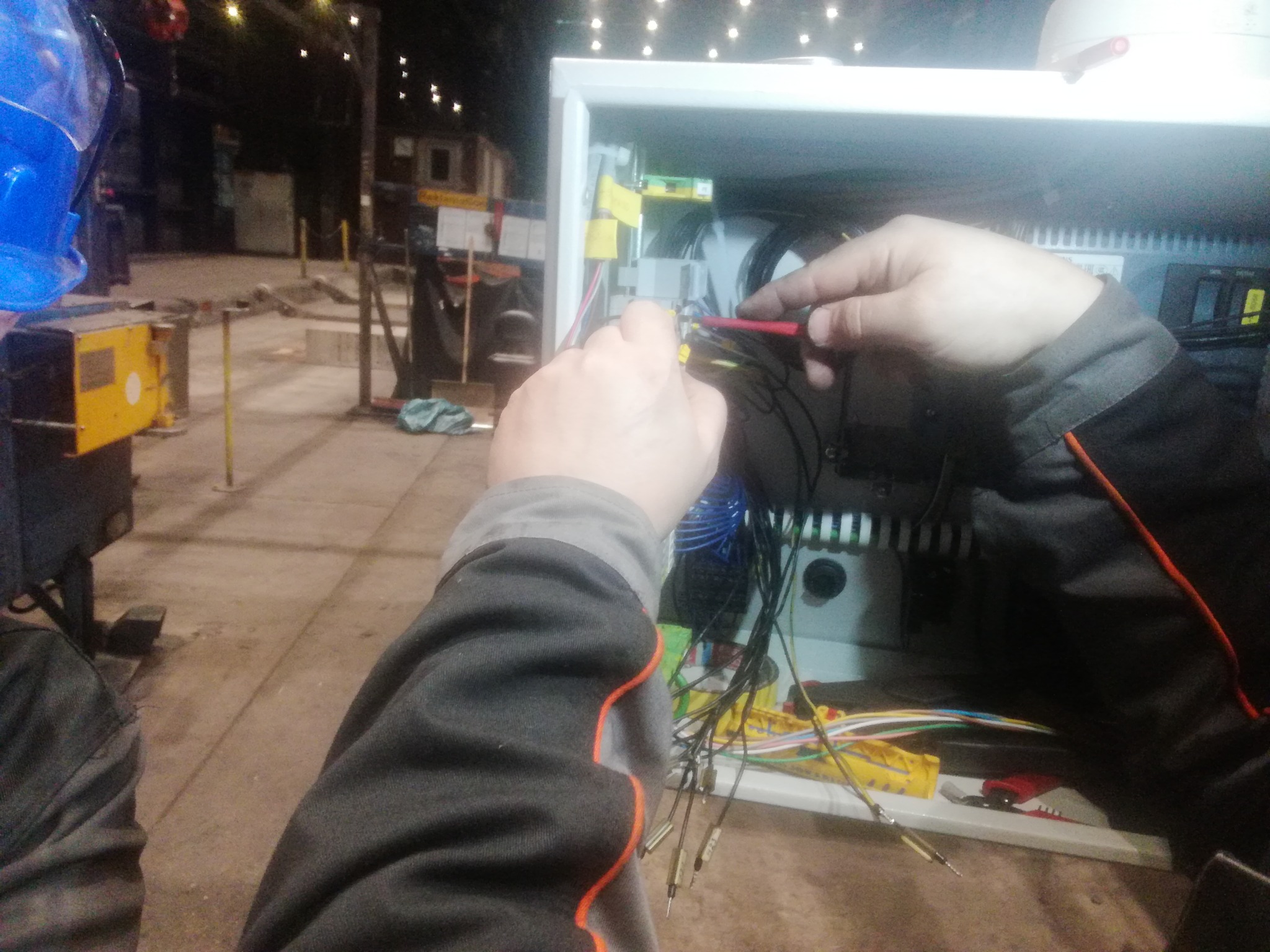 Elektryk mierzy napięcie w skrzynce elektrycznej za pomocą próbnika, widoczne przewody z końcówkami, wnętrze szafy sterowniczej i fragment ubrania osoby wykonującej pomiar.