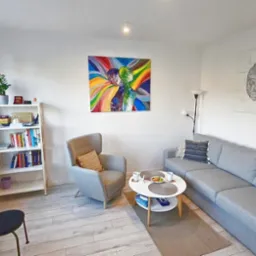 Jasne wnętrze gabinetu z szarą sofą, fotelem, stolikiem kawowym i regałem z książkami. Na ścianach wiszą obrazy o tematyce psychologicznej.