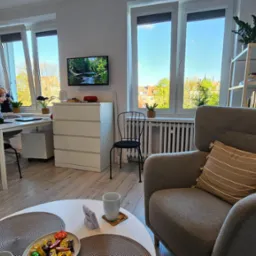 Jasny gabinet z biurkiem, krzesłem, fotelem i stolikiem kawowym. Na ścianie obraz i telewizor. W tle okno z widokiem na zieleń. Wnętrze urządzone w neutralnych barwach.