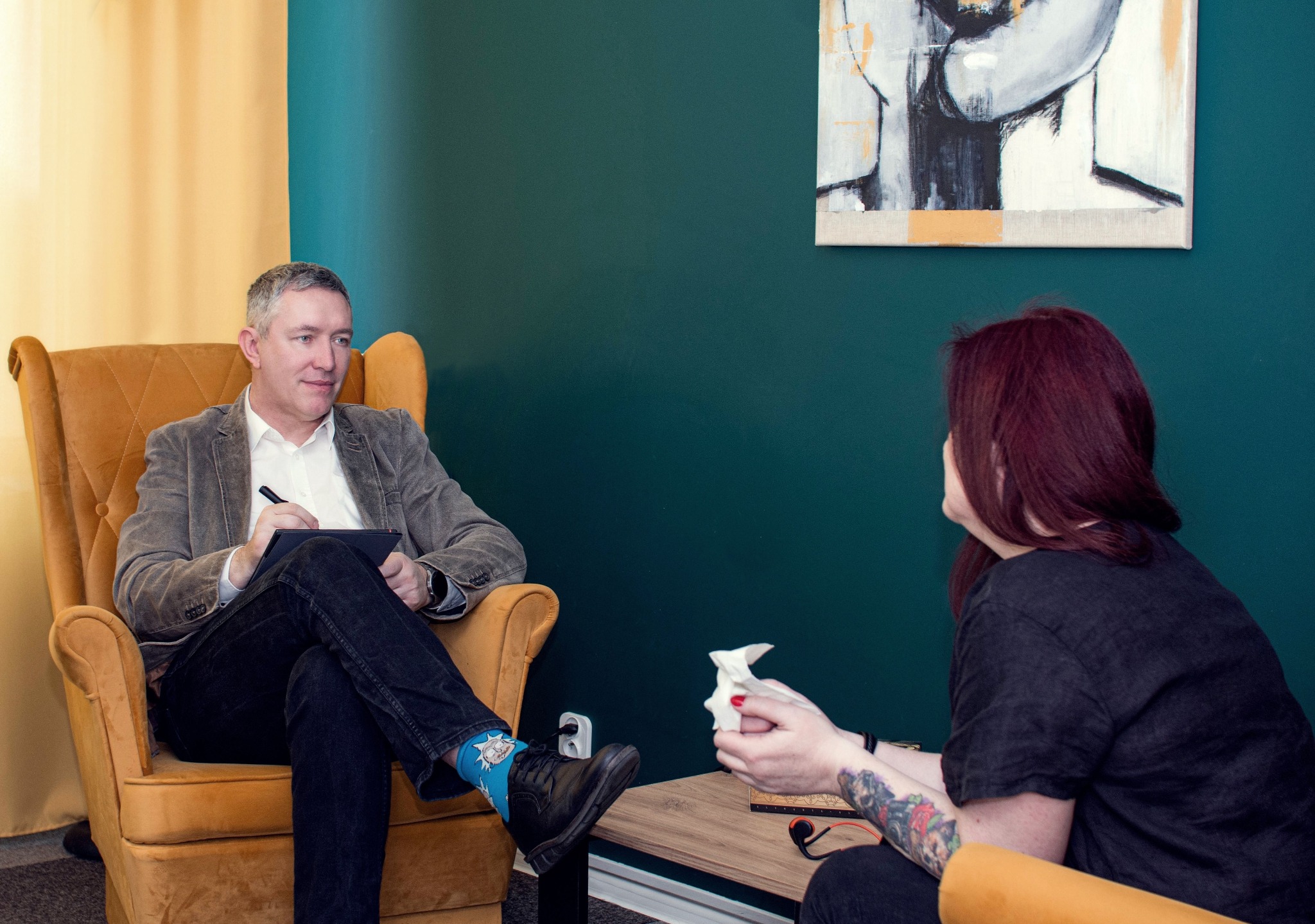 Mężczyzna w fotelu notuje na tablecie podczas sesji z kobietą trzymającą chusteczki, na ścianie abstrakcyjny obraz, mężczyzna ma na sobie skarpetki z motywem Ricka i Morty'ego.