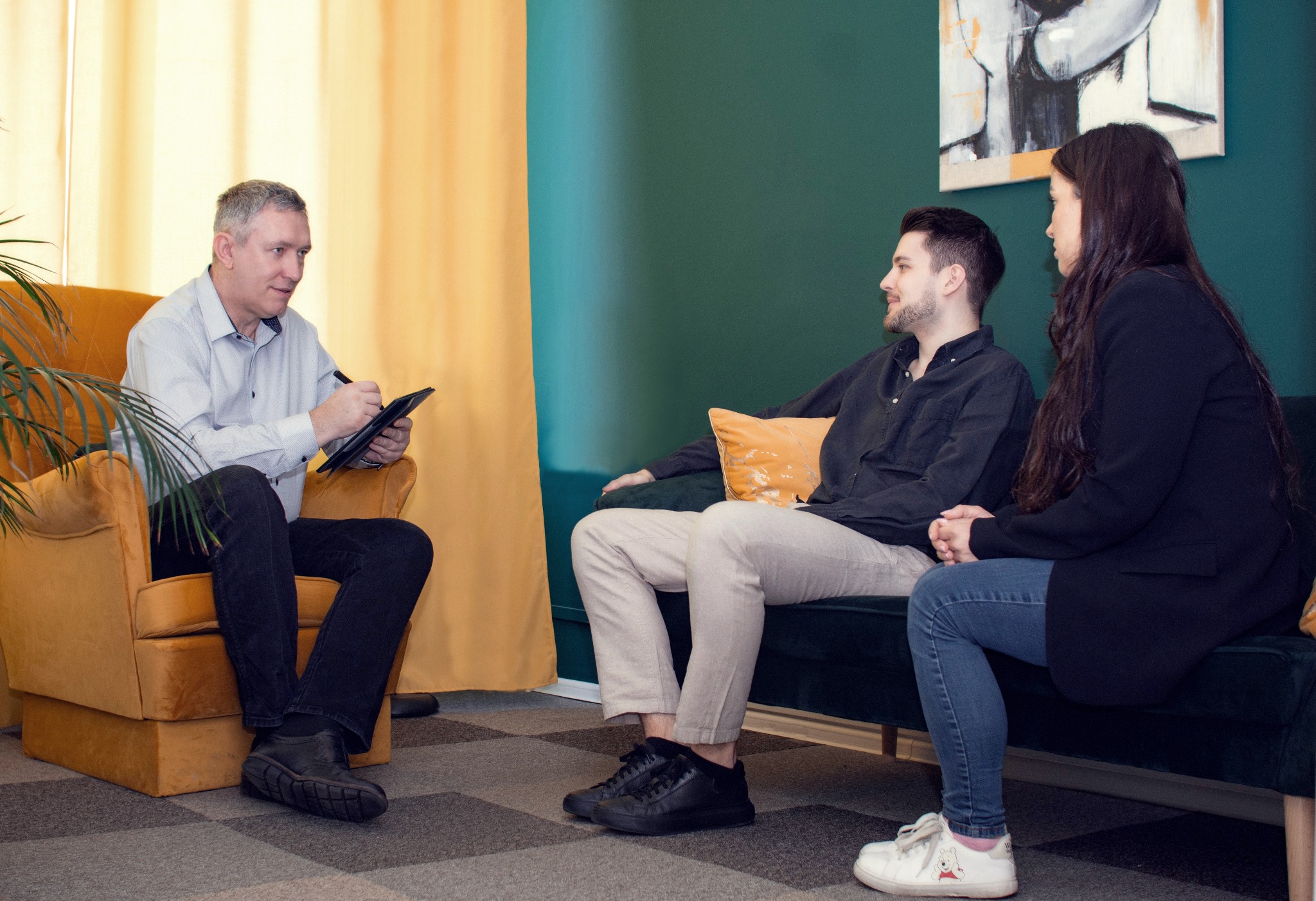 Mężczyzna w jasnej koszuli siedzi w pomarańczowym fotelu i robi notatki na tablecie, naprzeciwko niego siedzą na zielonej sofie młody mężczyzna i kobieta podczas konsultacji.