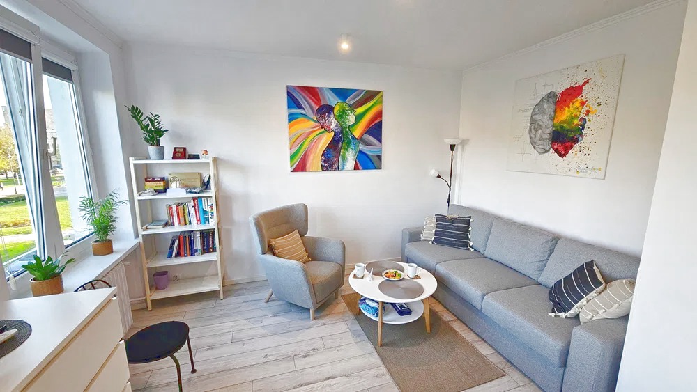 Jasne wnętrze gabinetu z szarą sofą, fotelem, stolikiem kawowym i regałem z książkami. Na ścianach wiszą obrazy o tematyce psychologicznej.