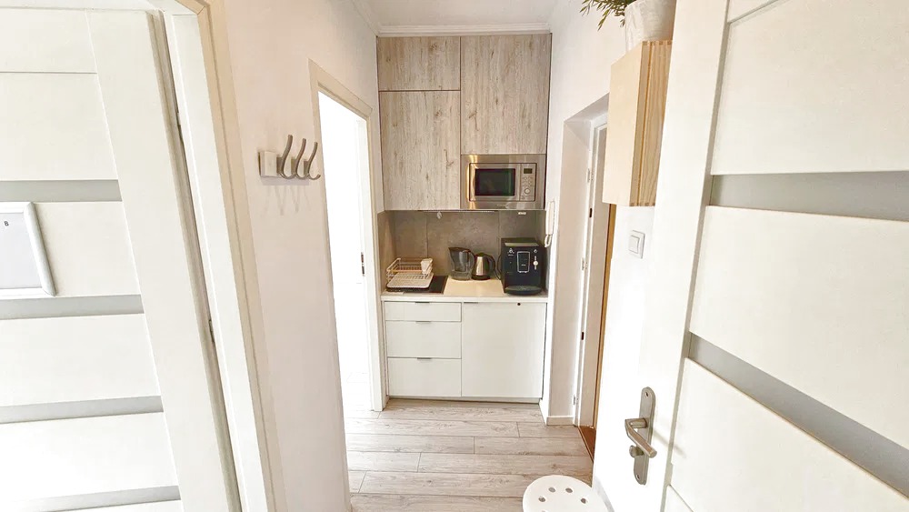 Nowoczesna, jasna kuchnia z drewnianymi szafkami, sprzętem AGD i białymi frontami. Widok z korytarza, z fragmentem białego krzesła barowego w dolnym rogu.