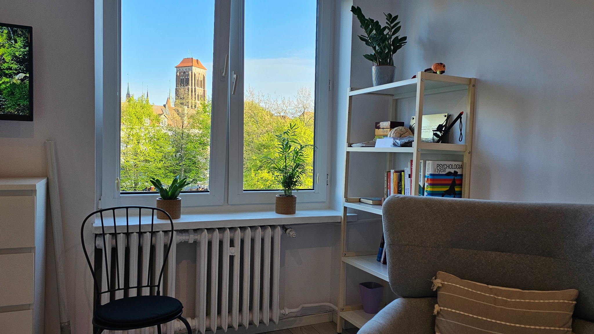 Pokój z oknem na zieleń i kościół. Na parapecie doniczki, obok regał z książkami, w tym 'Psychologia i życie'. Fotel z poduszką w rogu. Widok z gabinetu.