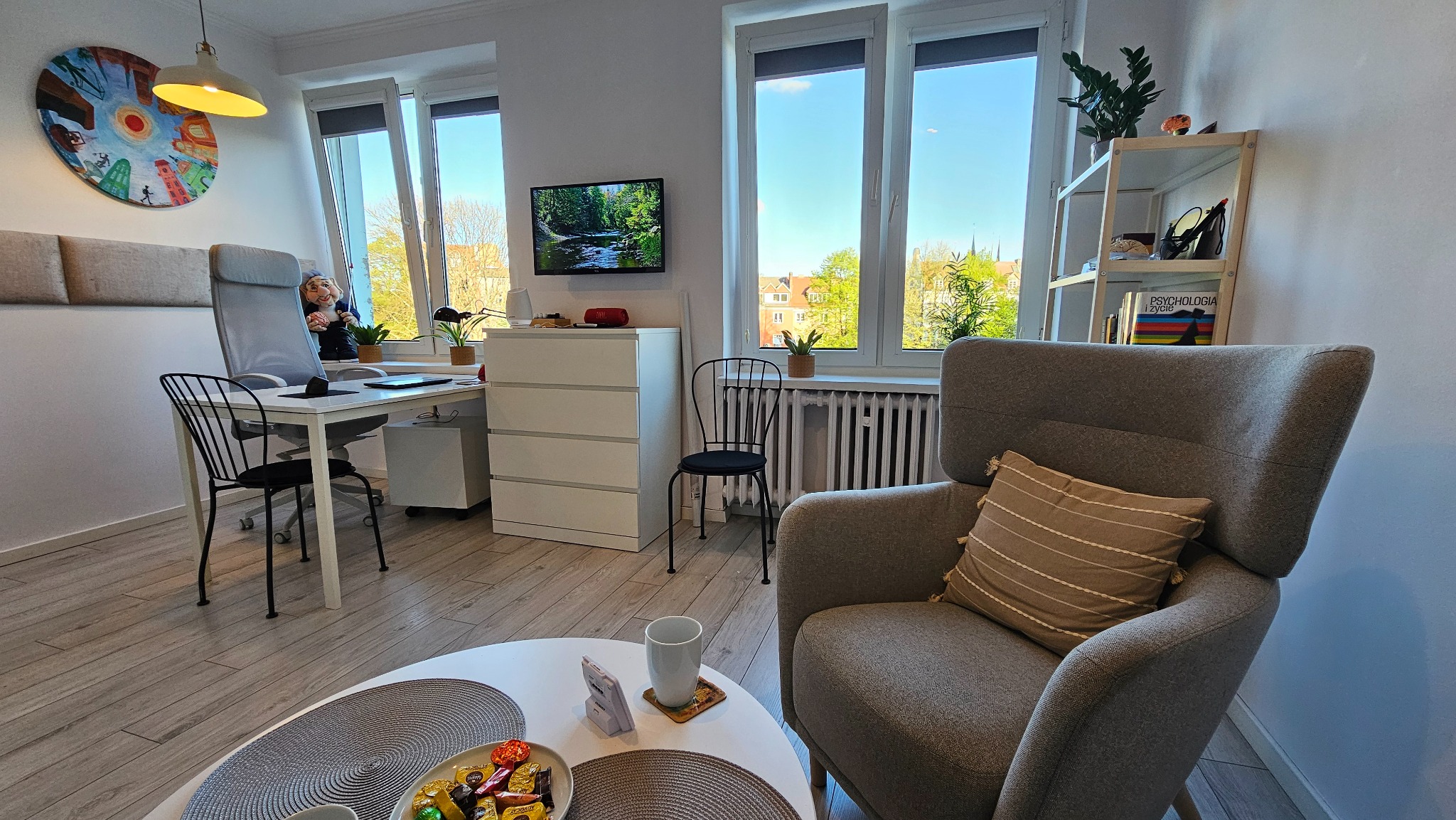 Jasny gabinet z biurkiem, krzesłem, fotelem i stolikiem kawowym. Na ścianie obraz i telewizor. W tle okno z widokiem na zieleń. Wnętrze urządzone w neutralnych barwach.