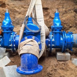 Solidna Ekipa - Instalacja wodociągowa z niebieskimi zaworami w wykopie, zabezpieczona pasami transportowymi, monter w pomarańczowych spodniach roboczych przyłącza element. Widoczne cegły i narzędzia.