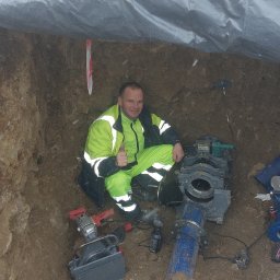 Solidna Ekipa - Pracownik w odblaskowym kombinezonie w wykopie, kciuk w górę, obok spawarka do rur, niebieska rura i narzędzia, ziemia pokryta śniegiem, wykop zabezpieczony plandeką.