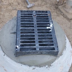 Solidna Ekipa - Studzienka kanalizacyjna z żeliwnym włazem osadzona w betonowym kręgu, obok leży młotek brukarski, widoczny ubijak wibracyjny.