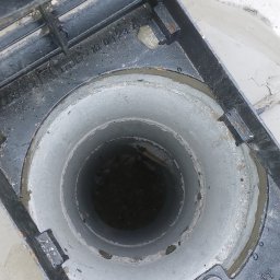 Solidna Ekipa - Otwarty właz betonowego zbiornika z widocznym wnętrzem studzienki i częściowo odsłoniętą pokrywą z metalową kratką, na tle ziemi.