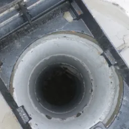 Otwarty właz betonowego zbiornika z widocznym wnętrzem studzienki i częściowo odsłoniętą pokrywą z metalową kratką, na tle ziemi.