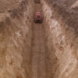Solidna Ekipa - Wykopany głęboki rów w ziemi z widoczną pomarańczową zagęszczarką w oddali, na górze widoczne drzewa i pochmurne niebo.