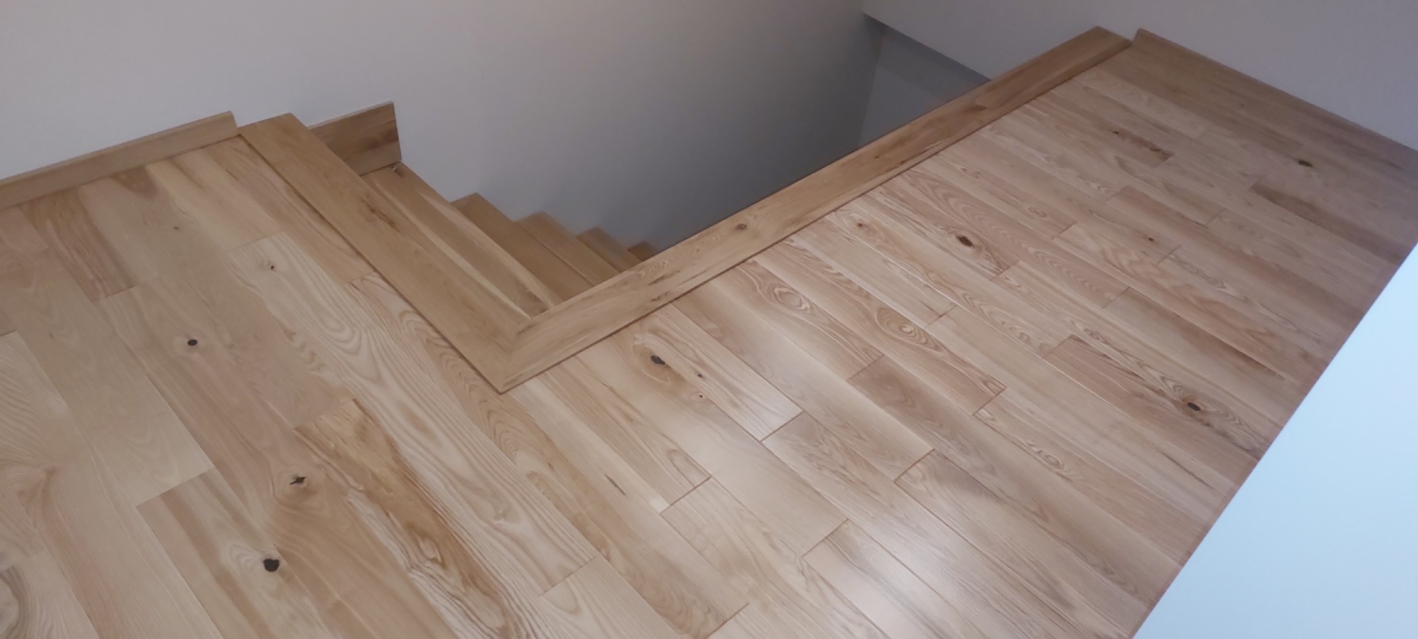 Jasne, drewniane panele podłogowe ułożone wzdłuż ściany i schodów; widoczne naturalne słoje i sęki drewna.