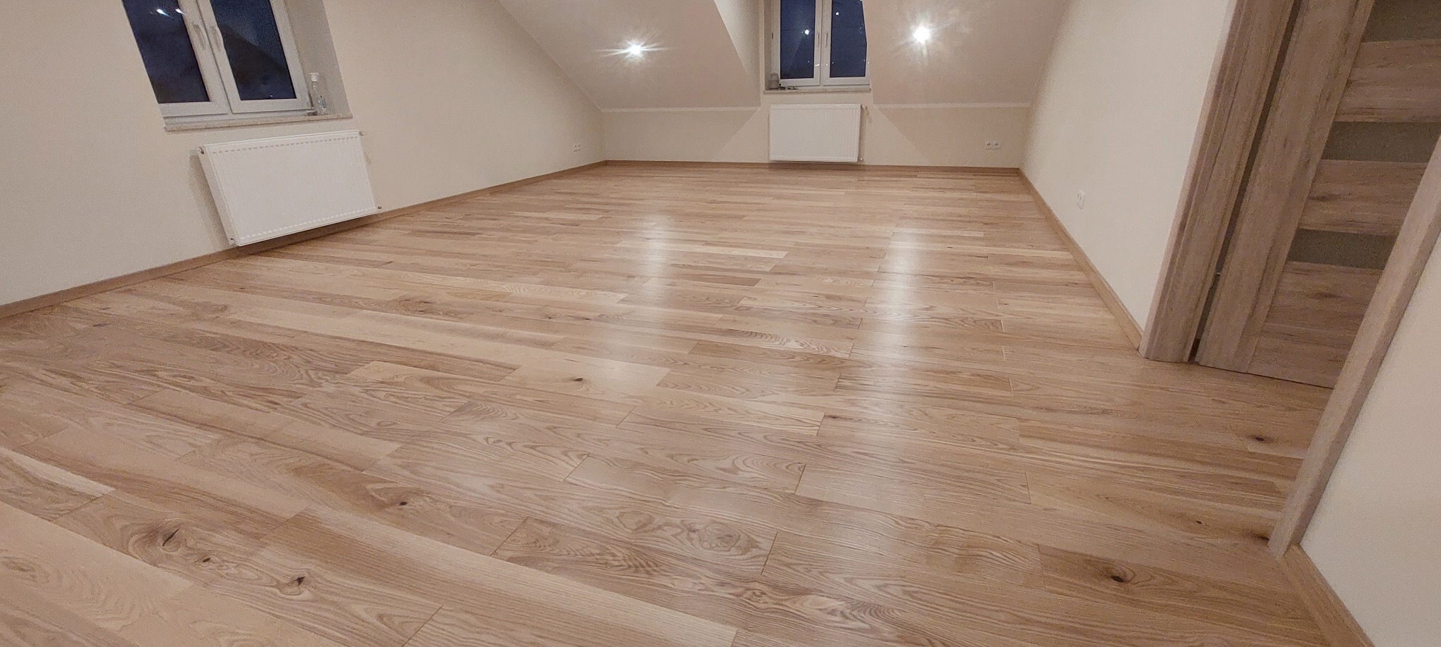 Jasne, drewniane panele podłogowe ułożone w przestronnym, jasnym pokoju na poddaszu, z widocznymi grzejnikami pod oknami i drewnianymi drzwiami w tle.