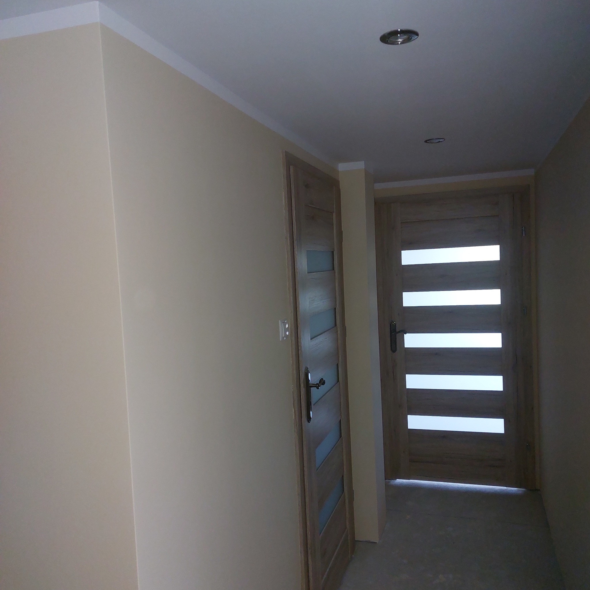 Korytarz po remoncie z dwiema drewnianymi drzwiami z szybami, pomalowane ściany w odcieniach beżu i szarości.