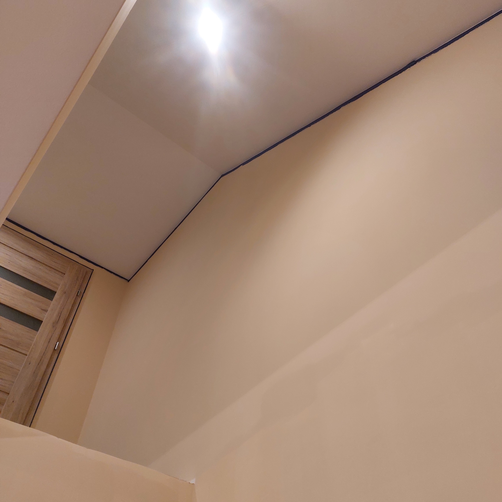 Pomieszczenie w trakcie malowania, widoczna ściana w odcieniach beżu, sufit z widocznym odcięciem koloru i fragment stolarki drzwiowej po lewej stronie.