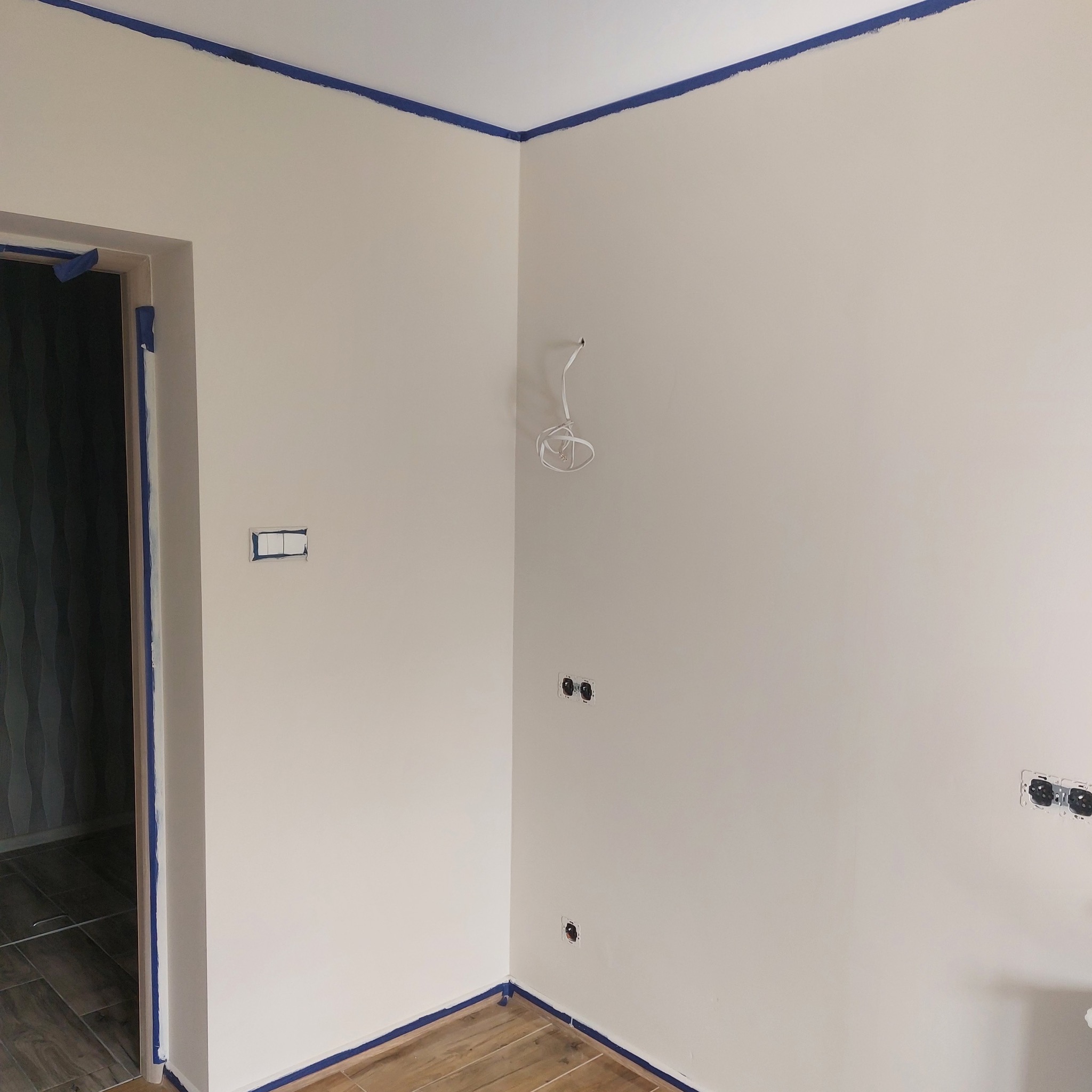 Pokój w trakcie malowania, zabezpieczone taśmą malarską krawędzie ścian, sufitu i futryny drzwi, widoczne puszki elektryczne bez osprzętu oraz wystające przewody.