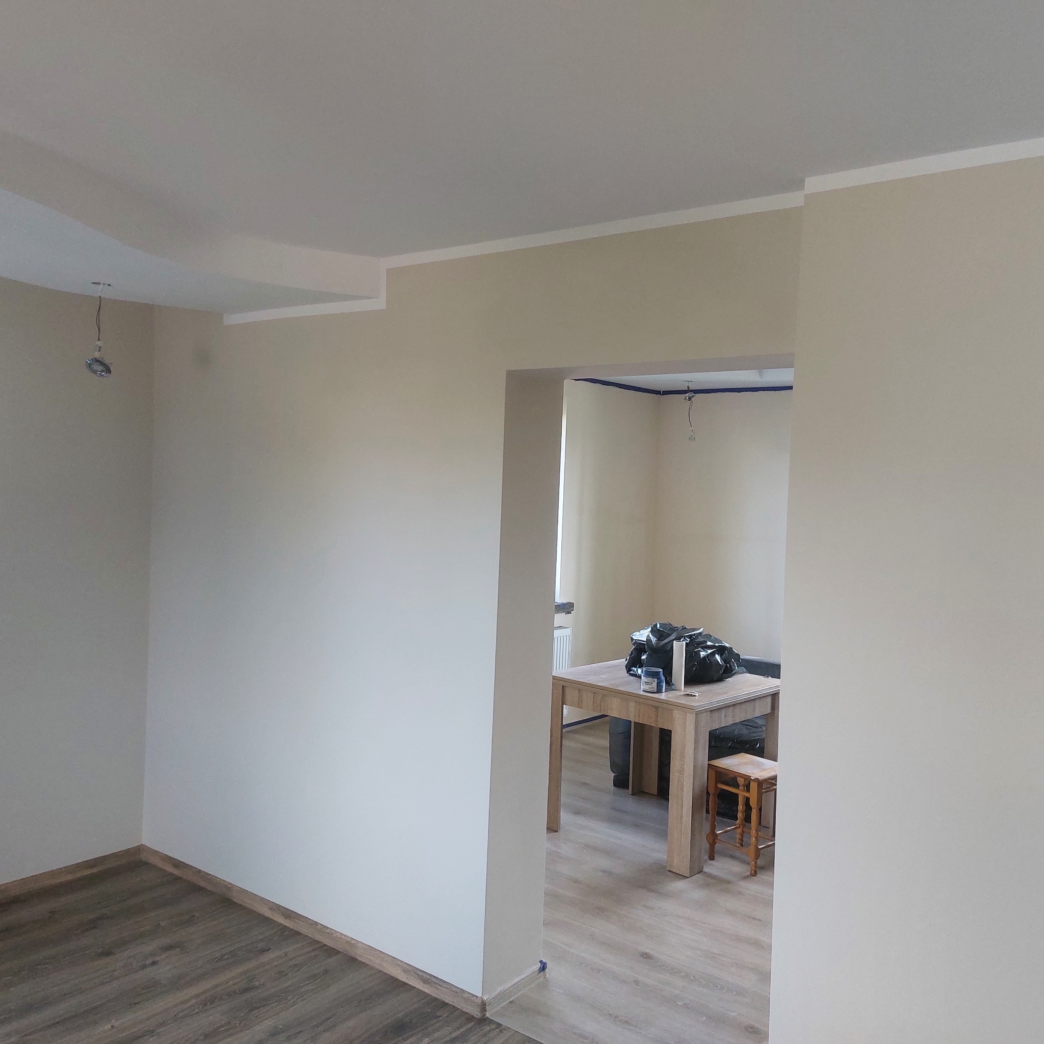 Wnętrze w trakcie remontu: świeżo pomalowane ściany w odcieniach beżu, białe listwy przysufitowe, drewnopodobne panele podłogowe, instalacja oświetleniowa bez opraw, widok na sąsiednie...