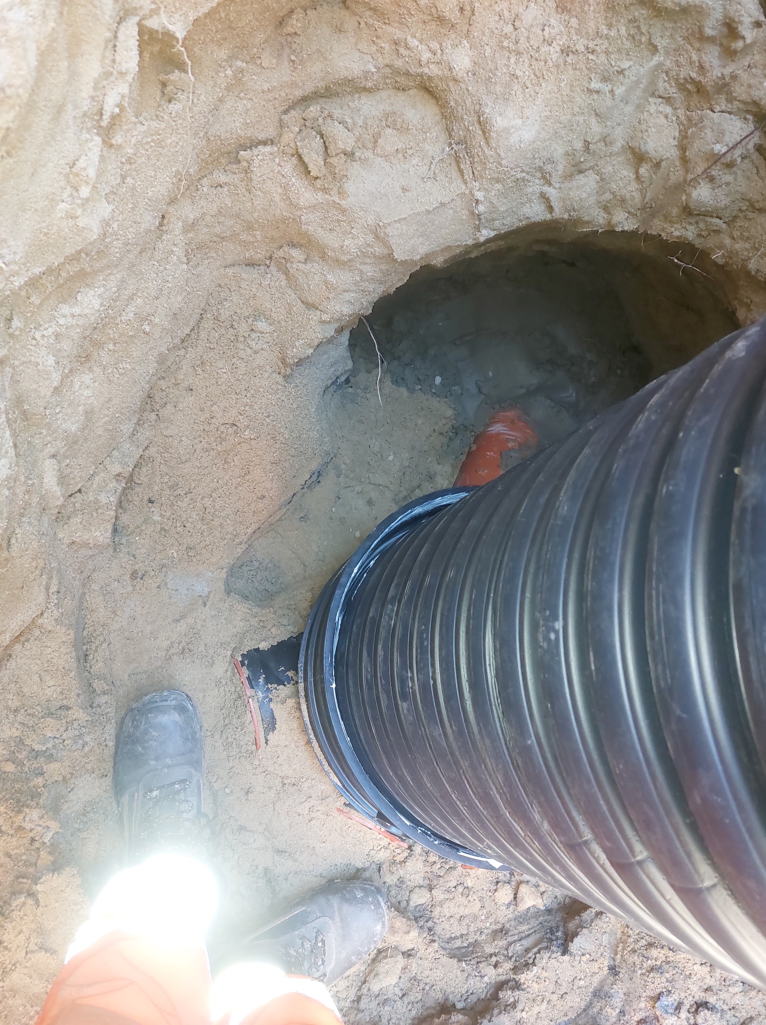 Czarny karbowany rurociąg w wykopie z piaszczystą ziemią, widoczne buty robocze w dolnej części kadru, pomarańczowy element rury w głębi wykopu.