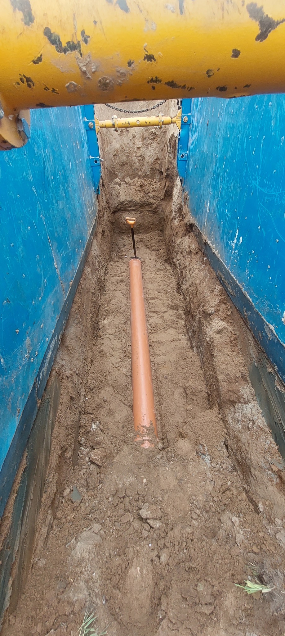 Pomarańczowa rura ułożona w wykopie zabezpieczonym niebieską szalunką, widoczna łopata, prace ziemne w Garbowie.
