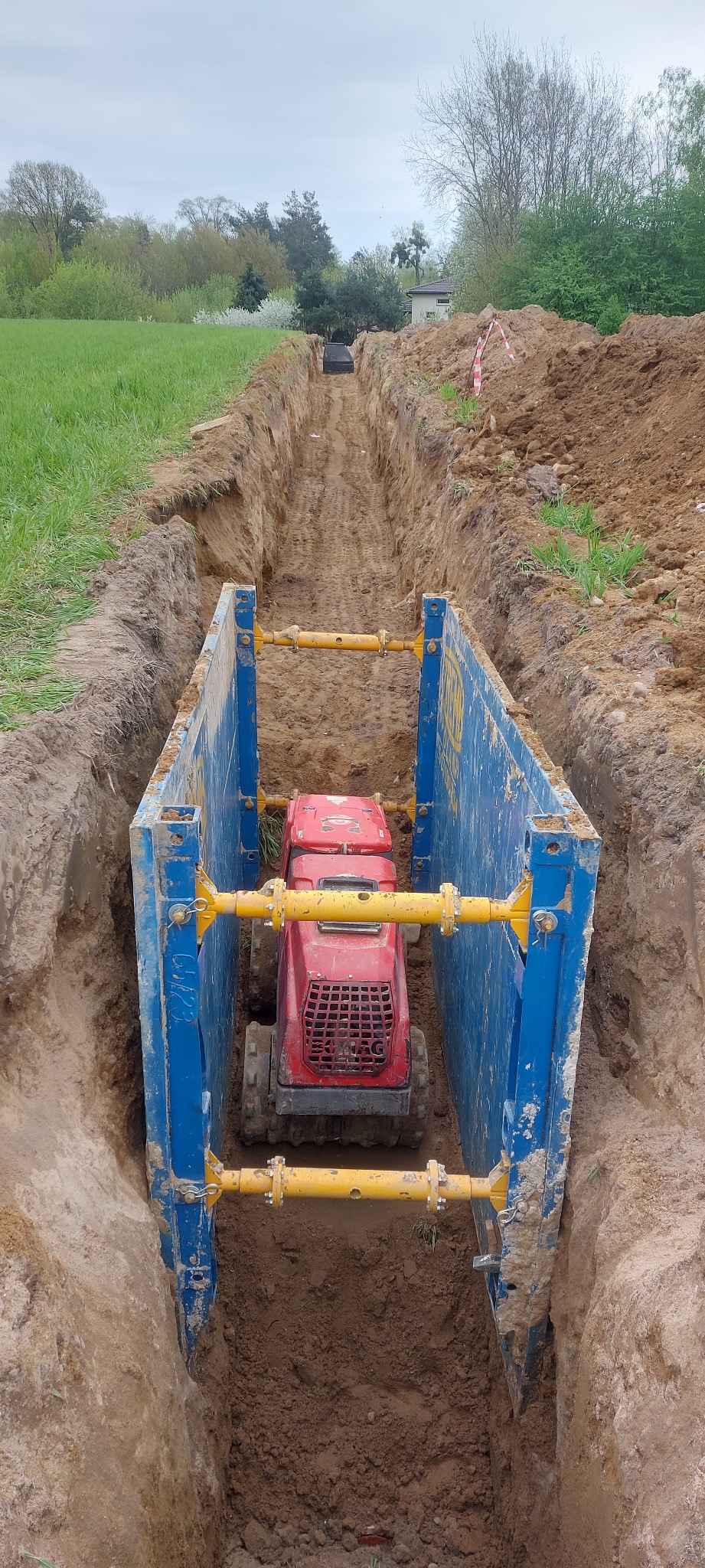 Czerwona zagęszczarka gruntu w wykopie zabezpieczonym niebieskimi szalunkami, widok na zielone pole i drzewa w tle.