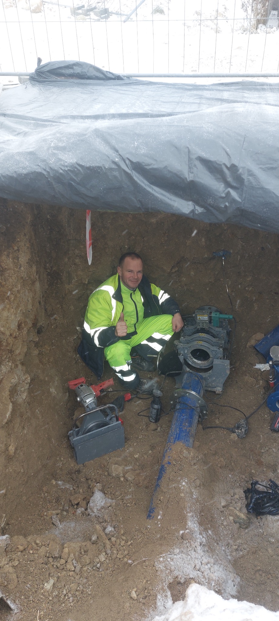 Pracownik w odblaskowym kombinezonie w wykopie, kciuk w górę, obok spawarka do rur, niebieska rura i narzędzia, ziemia pokryta śniegiem, wykop zabezpieczony plandeką.