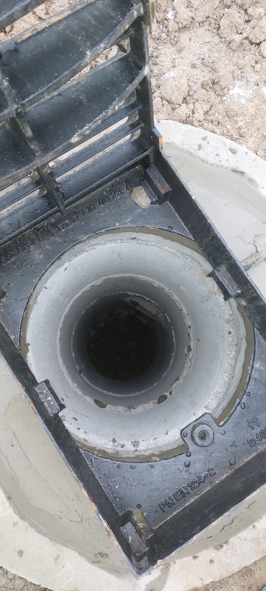 Otwarty właz betonowego zbiornika z widocznym wnętrzem studzienki i częściowo odsłoniętą pokrywą z metalową kratką, na tle ziemi.