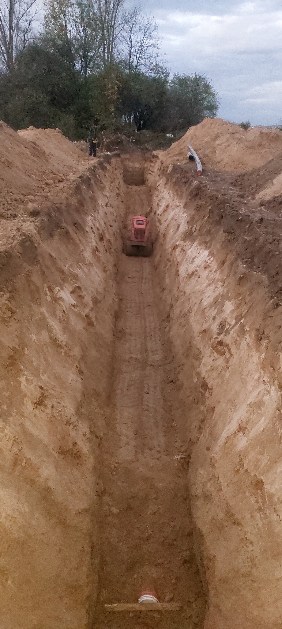 Wykopany głęboki rów w ziemi z widoczną pomarańczową zagęszczarką w oddali, na górze widoczne drzewa i pochmurne niebo.