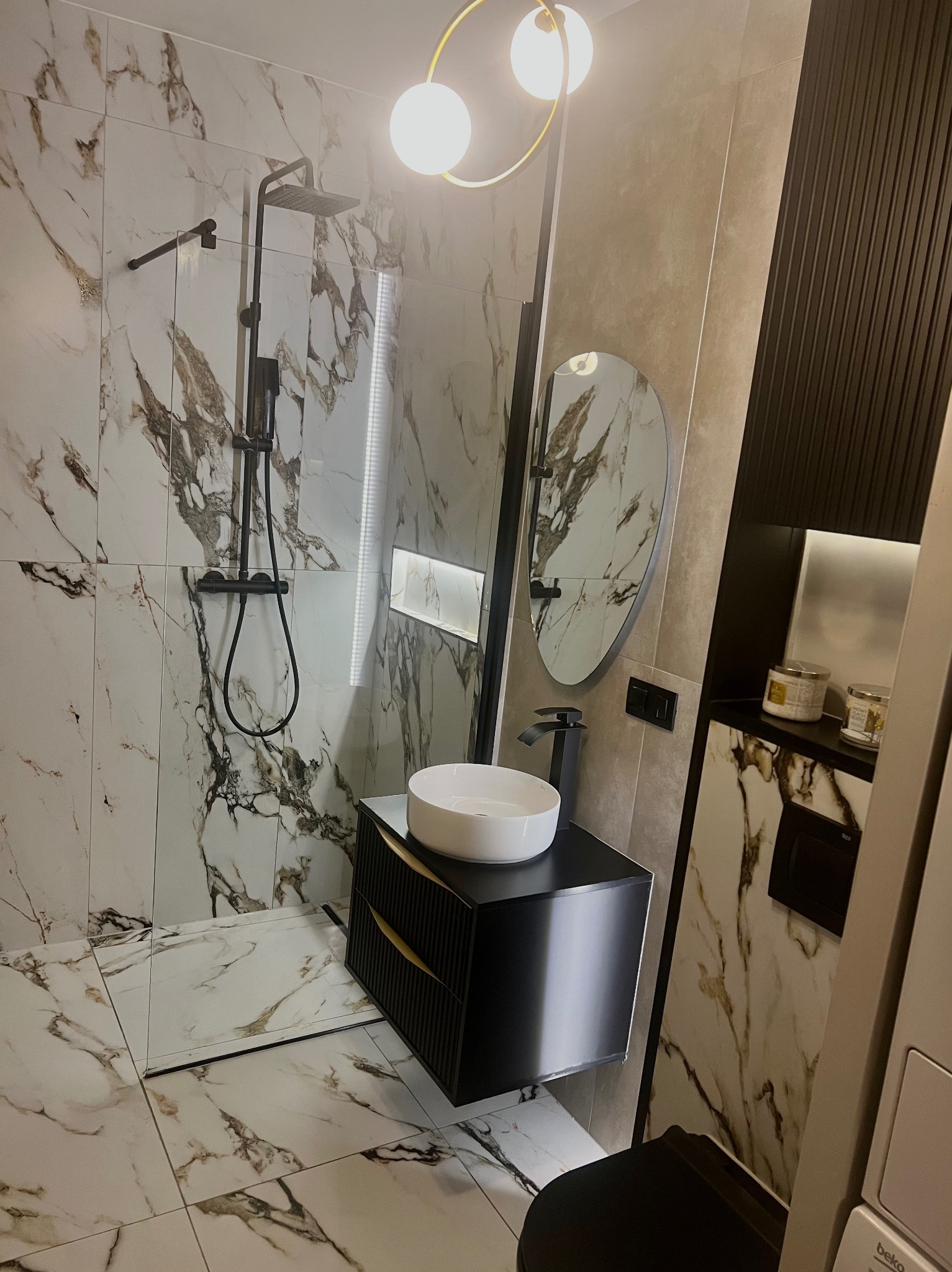 Nowoczesna łazienka z kabiną prysznicową typu walk-in, umywalką nablatową na czarnej szafce z frezowanymi frontami, okrągłym lustrem i marmurowymi płytkami na ścianach i podłodze.