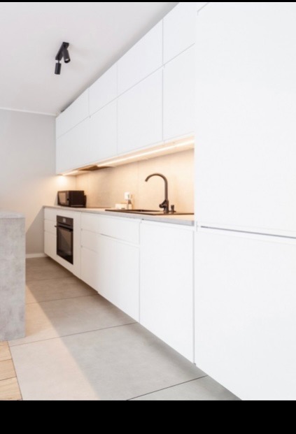 Minimalistyczna kuchnia z białymi, gładkimi frontami szafek, czarną armaturą i oświetleniem LED pod szafkami wiszącymi.