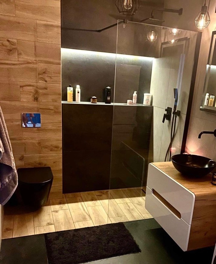 Nowoczesna łazienka z czarną armaturą, drewnopodobnymi płytkami i oświetleniem LED, widoczna kabina prysznicowa typu walk-in oraz umywalka nablatowa.