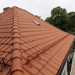 Artbudownictwo | Dachy | Elewacje | Słupsk | - Czerwona dachówka ceramiczna na skośnym dachu z dwoma oknami dachowymi i zabezpieczeniem przeciwśniegowym, widoczny komin i rusztowanie.