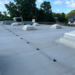 Artbudownictwo | Dachy | Elewacje | Słupsk | - Szary dach przemysłowy z świetlikami kopułowymi i systemem zabezpieczeń linowych, pracownik w odblaskowej kamizelce w oddali, budynki i drzewa w tle.