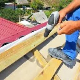 Artbudownictwo | Dachy | Elewacje | Słupsk | - Pracownik w niebieskich spodniach przykręca drewnianą łatę do konstrukcji dachu pokrytego czerwoną blachodachówką za pomocą wkrętarki akumulatorowej, widok z góry.