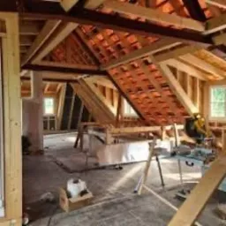 Poddasze w trakcie adaptacji: widoczne drewniane elementy konstrukcyjne dachu, krokwie, izolacja między nimi, okna w ścianie szczytowej, narzędzia budowlane na podłodze, częściowo oświetlone...