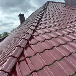 Błyszczący, bordowy dach z dachówki ceramicznej, widok z dołu na tle pochmurnego nieba, z kominem w tle.