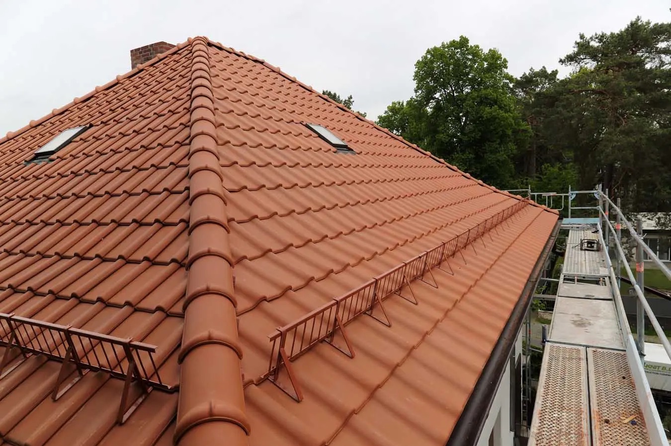 Czerwona dachówka ceramiczna na skośnym dachu z dwoma oknami dachowymi i zabezpieczeniem przeciwśniegowym, widoczny komin i rusztowanie.