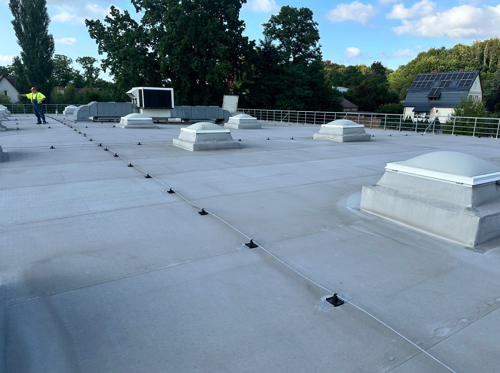 Szary dach przemysłowy z świetlikami kopułowymi i systemem zabezpieczeń linowych, pracownik w odblaskowej kamizelce w oddali, budynki i drzewa w tle.